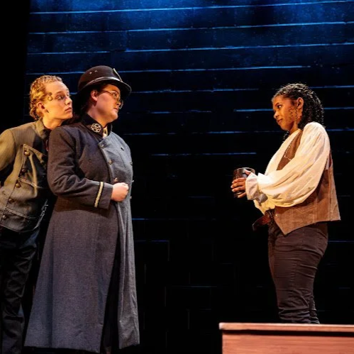 Heather is onstage in a blue wool coat and a Victorian sheriff's cap. She locks eyes with a Black actor wearing a brown suede vest and white overshirt, as a white actor in a grey buttoned coat and gears on their face leans over Heather's shoulder.