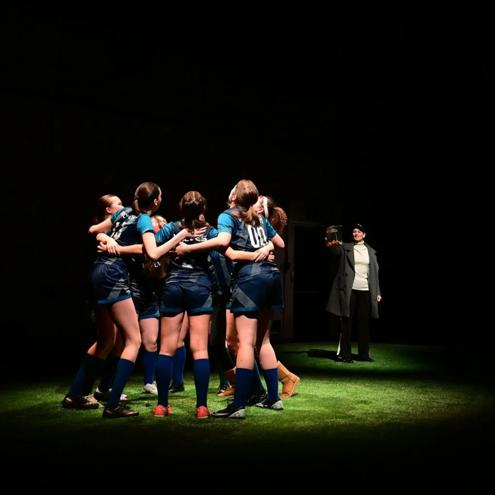 Young AFAB soccer players in dark blue uniforms stand on a pitch with shadowy lighting. They hold one another and look to the sky, as a middle-aged white woman stands behind with orange slices.