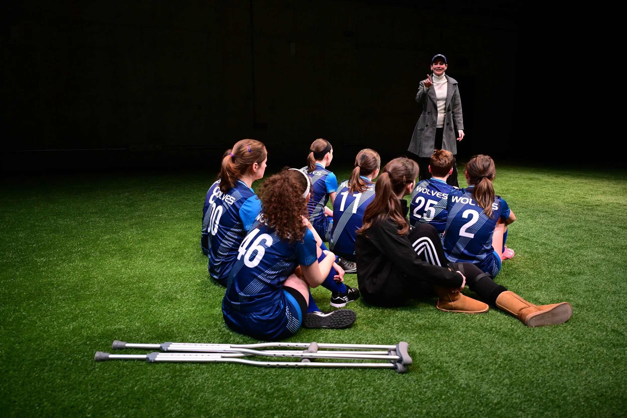 Young AFAB soccer players in dark blue uniforms sit on a pitch with shadowy lighting. They sit close to one another, listening to a middle-aged white woman in a peacoat.