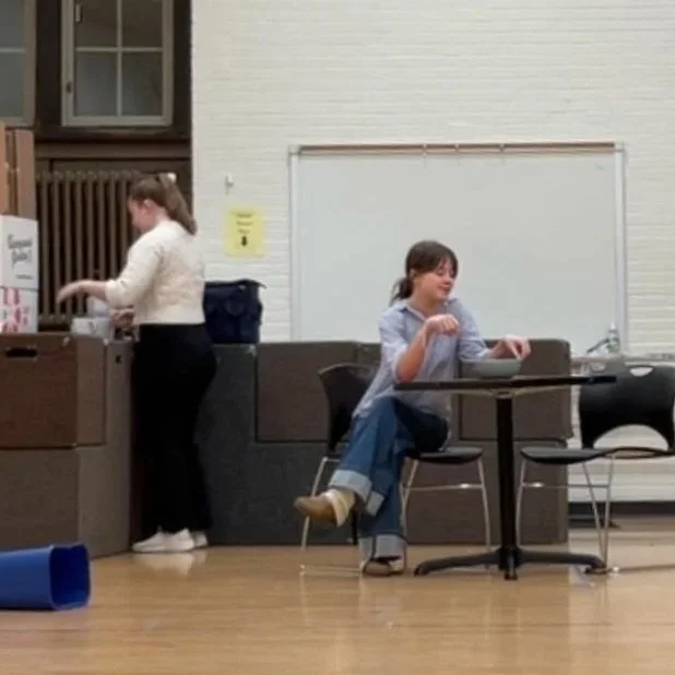 A college-age white woman in a blue striped shirt sits at a table. A college-age white woman in a white blouse stands near acting blocks made to look like a kitchen counter. They turn their backs to each other.