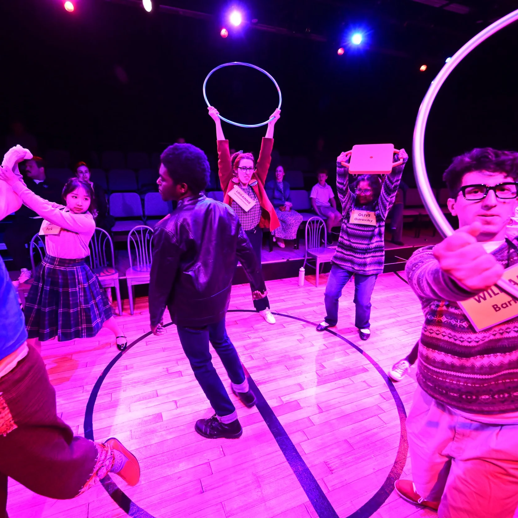 The cast dances and spins energetically, carrying hula hoops and scooters. The lighting is bright pink and vibrant.