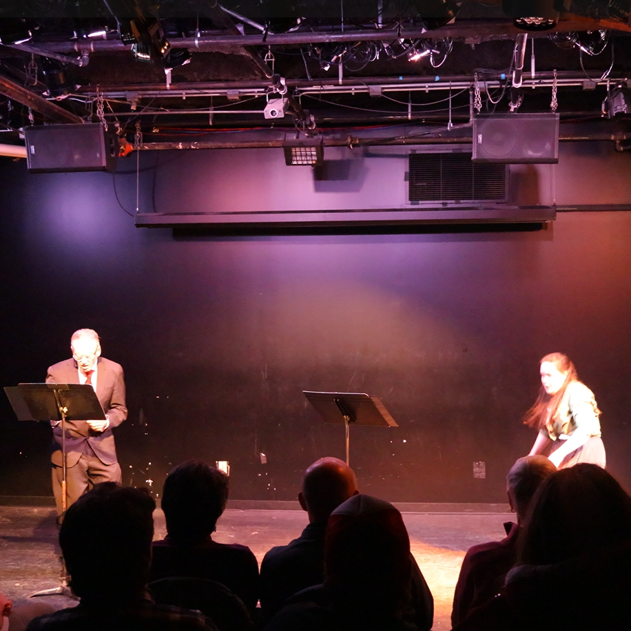 Heather bursts onstage, hunched over and panicked. She wears a green buttoned shirt with flowing black pants. A middle-aged white man in glasses and a blue suit looks at a music stand, bewildered.