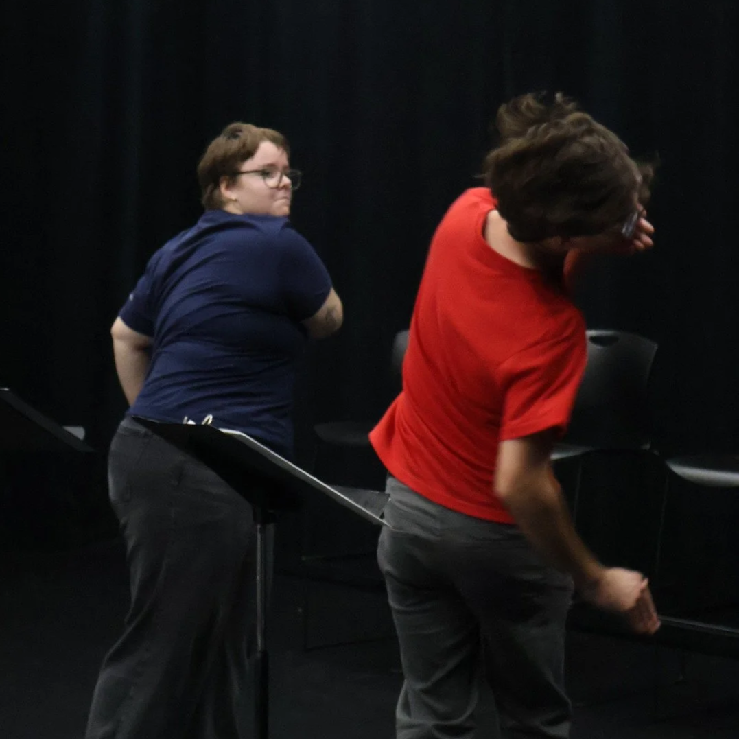 Two white college-age actors stand in front of music stands. An actor with glasses, short hair, and a blue shirt pretends to slap an actor with glasses, short dark hair, and a red shirt. The actor in the red shirt twists away, hair flying.