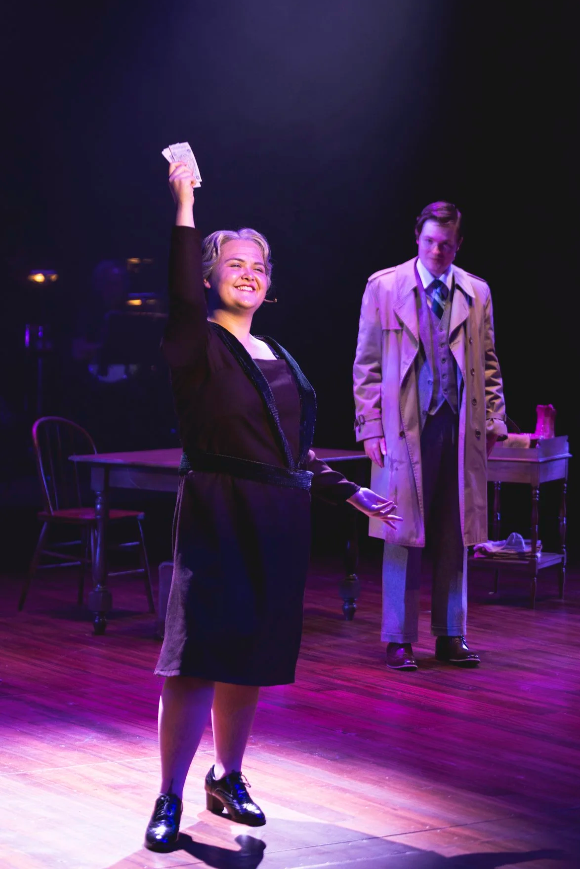 Heather is onstage, wearing a grey wig and a purple 1920s-style dress. She smiles wide, holding money in the air.