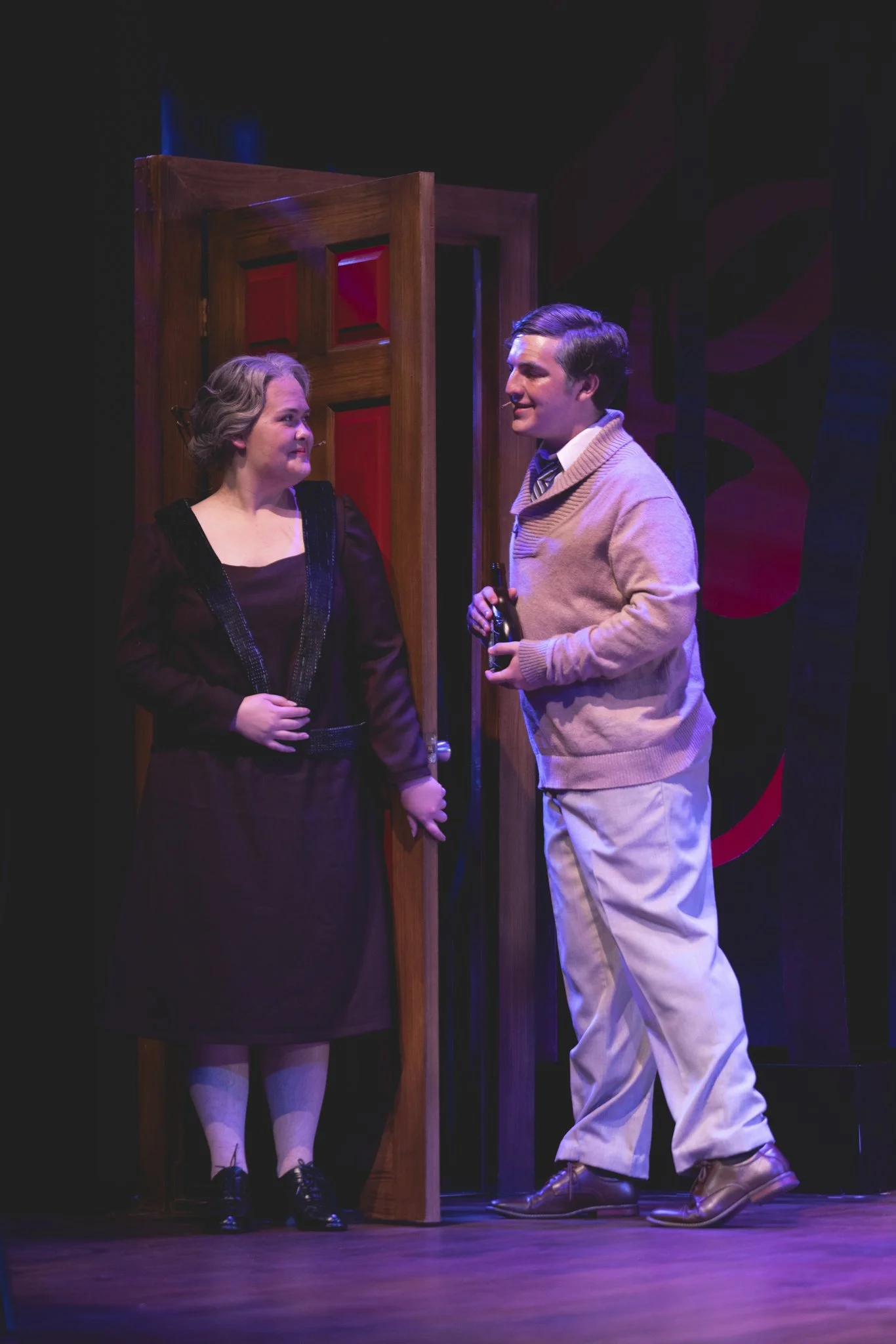 Heather is onstage in a grey wig and purple 1920s-style dress. She leans against a door, smiling coyly at a college-age man with light skin, greyed hair, and a beige sweater.