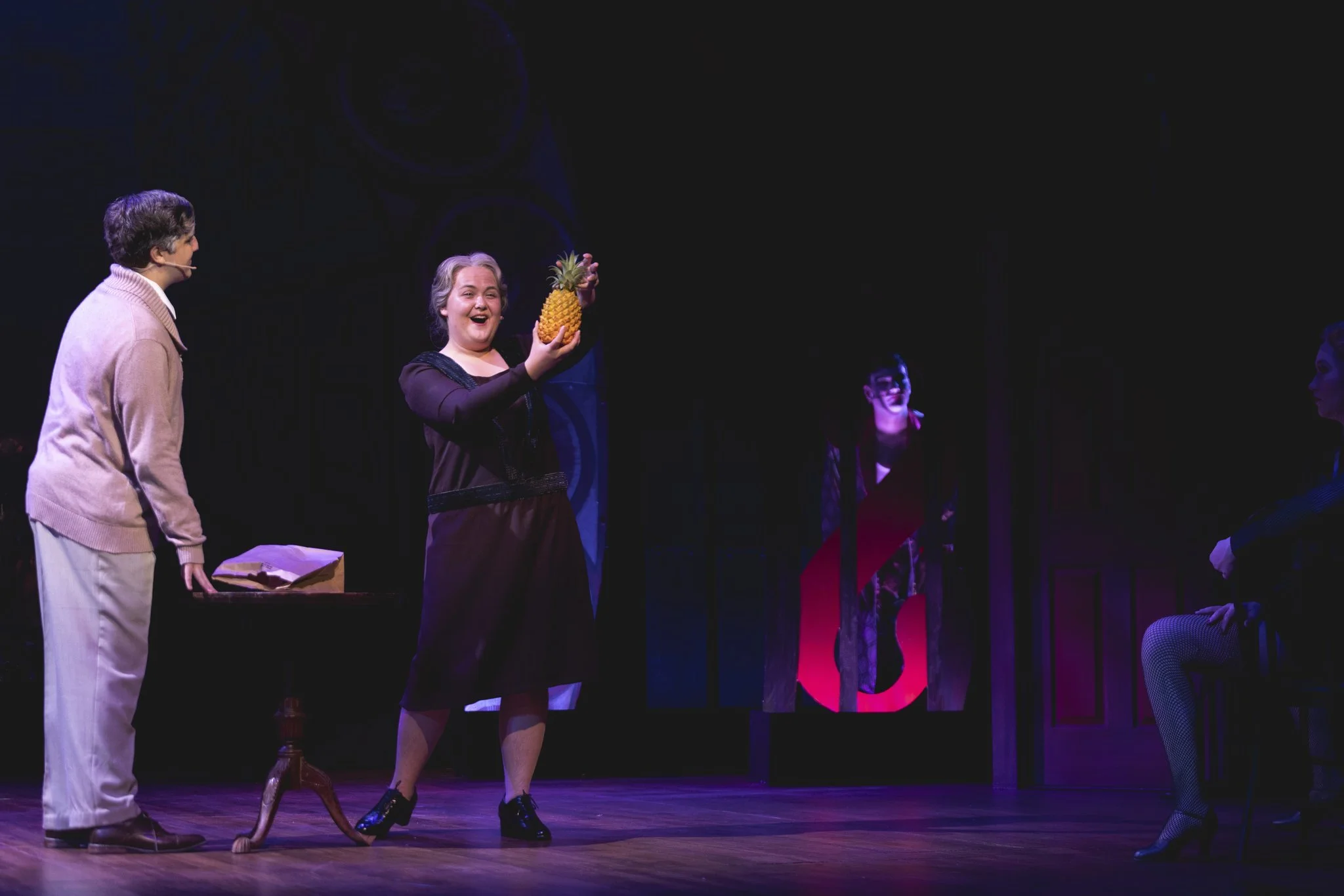 Heather is onstage in a grey wig and purple 1920s-style dress, gleefully holding a pineapple in the air. A college-age man in white face paint stands behind in red, ominous lighting.