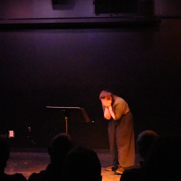 Heather is onstage beside a music stand. Her hair is half-up half-down, and she wears a green buttoned shirt with flowing black pants. Under dim orange lighting, she folds over, covering her face in grief.