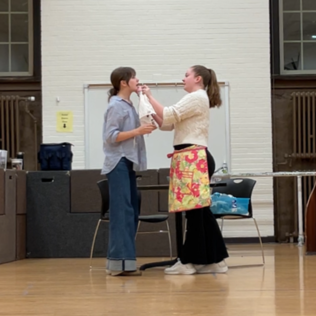 A college-age white woman in a pink flowered apron holds a rag to another college-age white woman's bloody nose and chin.