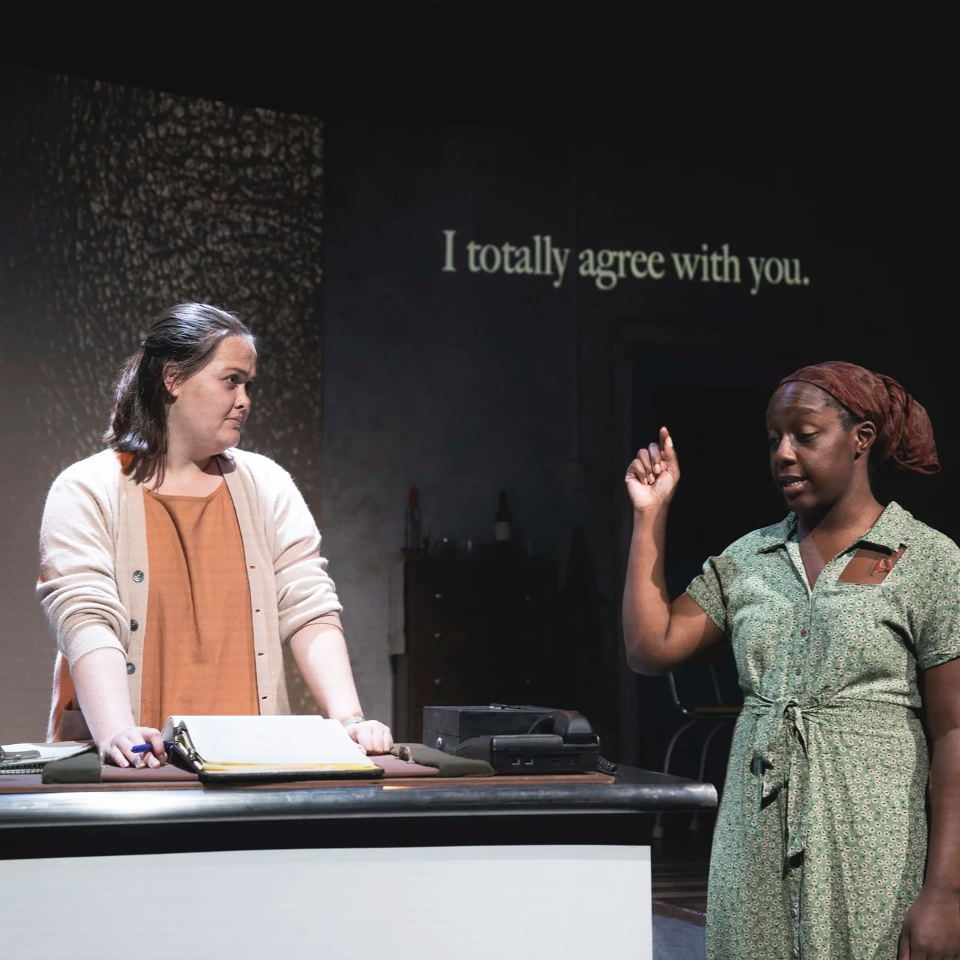 Heather is onstage in an orange dress and beige sweater. She listens skeptically to a Black woman in her 20s, who wears a brown hair wrap, a green dress, and an A-shaped brand on her chest. Projected text reads, "I totally agree with you."
