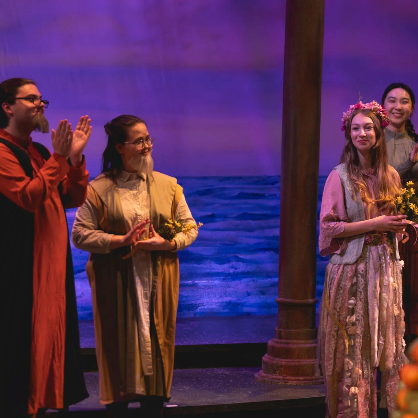 Heather is onstage, holding yellow flowers and standing in a crowd. She wears glasses, a rustic tan coat, a long white tunic, and a fake white beard. She claps with dignified approval.