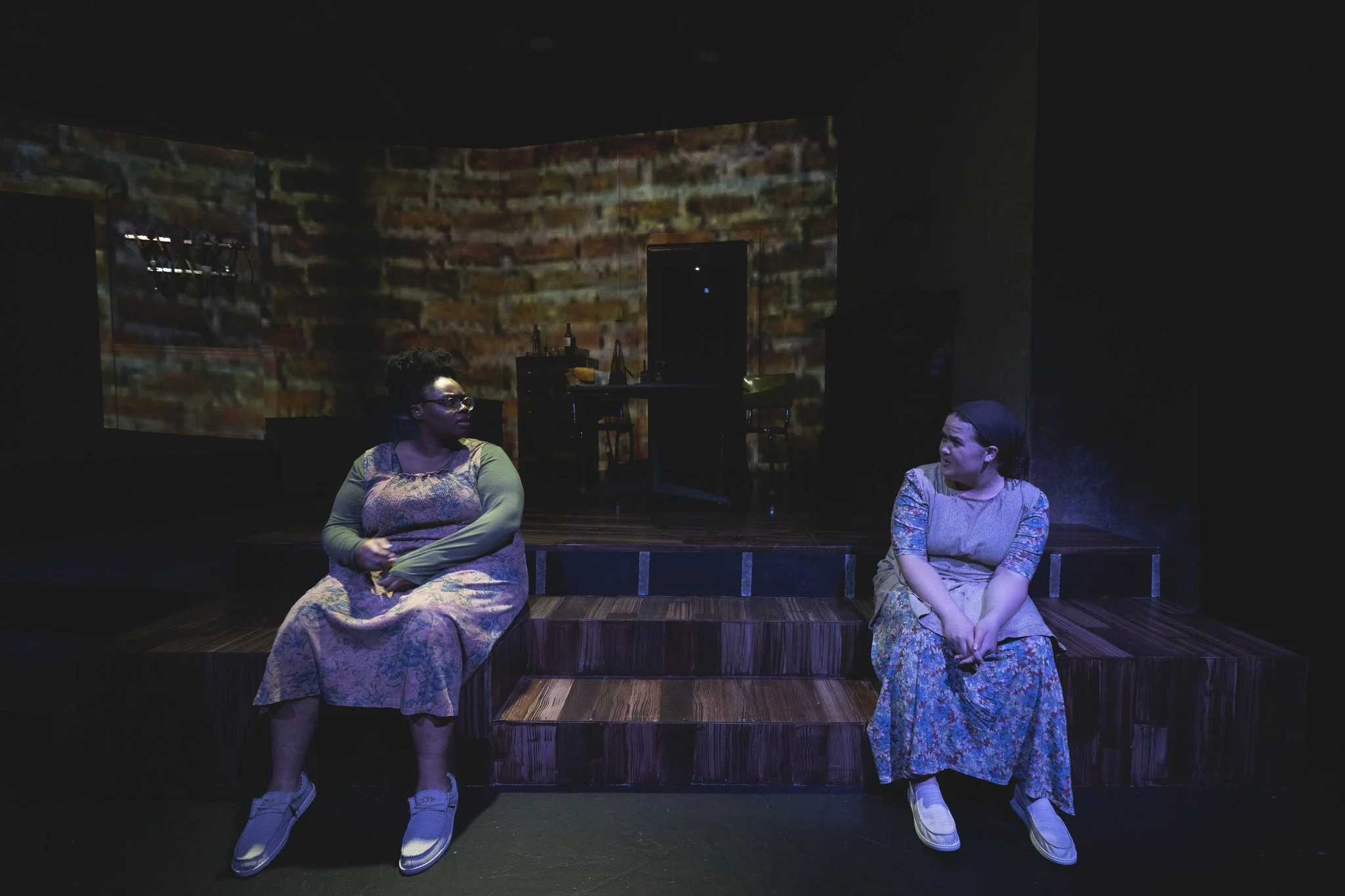 Heather is onstage in a faded blue floral dress, grey apron, and dark hair wrap. She sits on steps and speaks concernedly to a Black woman in her 20s, who wears glasses, a green shirt, and a faded pink floral dress.