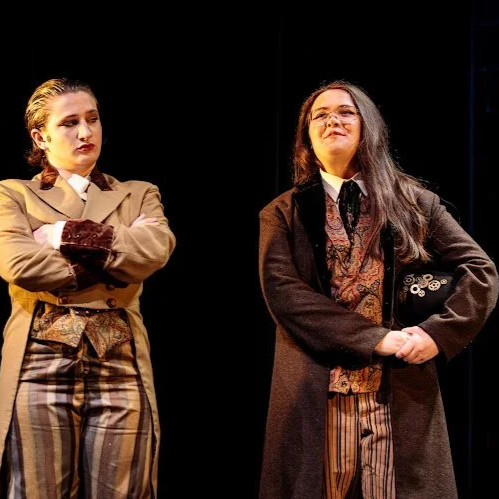 Heather is onstage in a brown wool peacoat and a patterned vest. She holds a black top hat with gears, and speaks reflectively. A white actor with slicked short hair and strong makeup crosses their arms as they listen.
