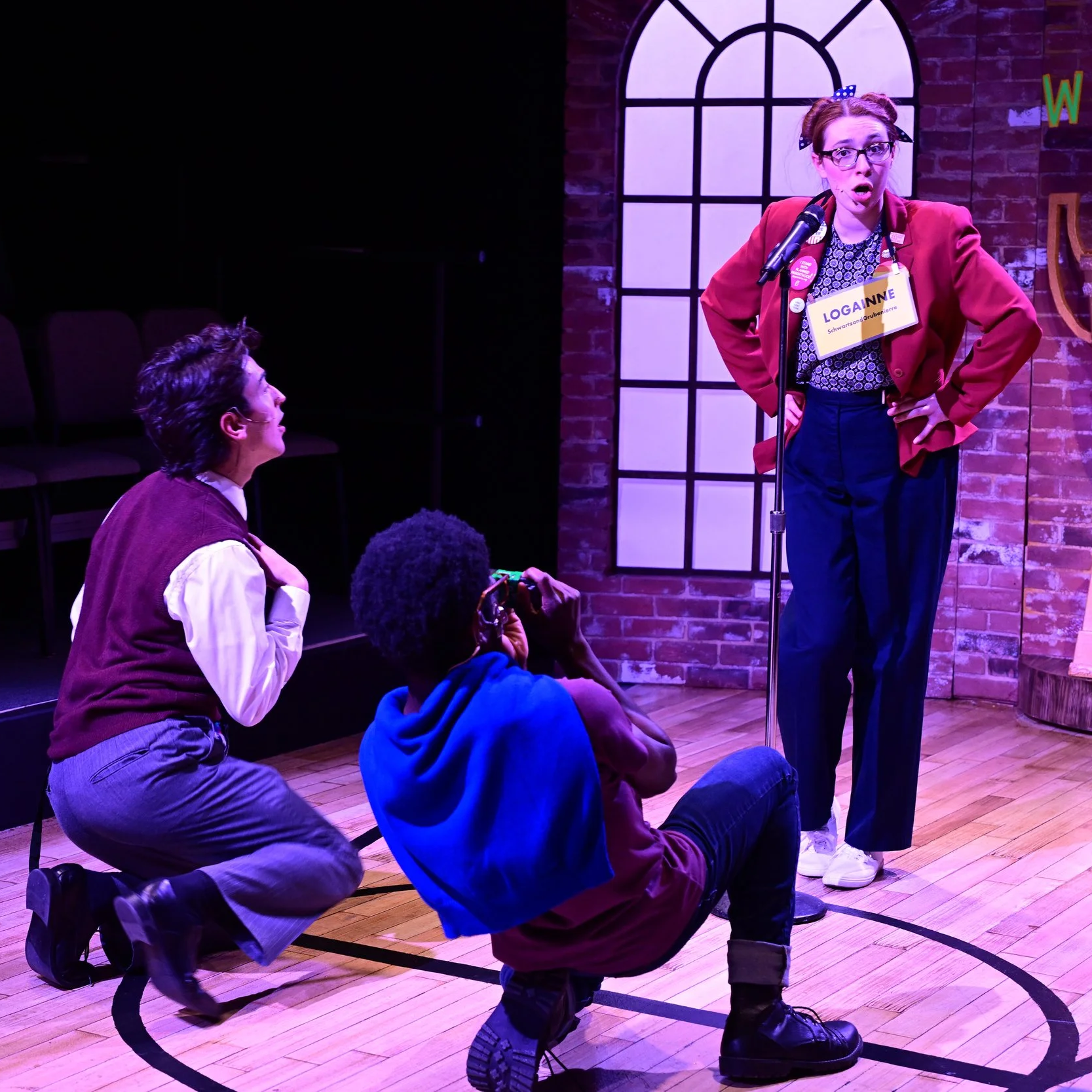 A college-age white woman stands in front of a microphone, wearing glasses and a red jacket covered with social justice pins. She sings, hands on her hips, as two college-age men kneel before her in paternal adoration. One takes her picture.