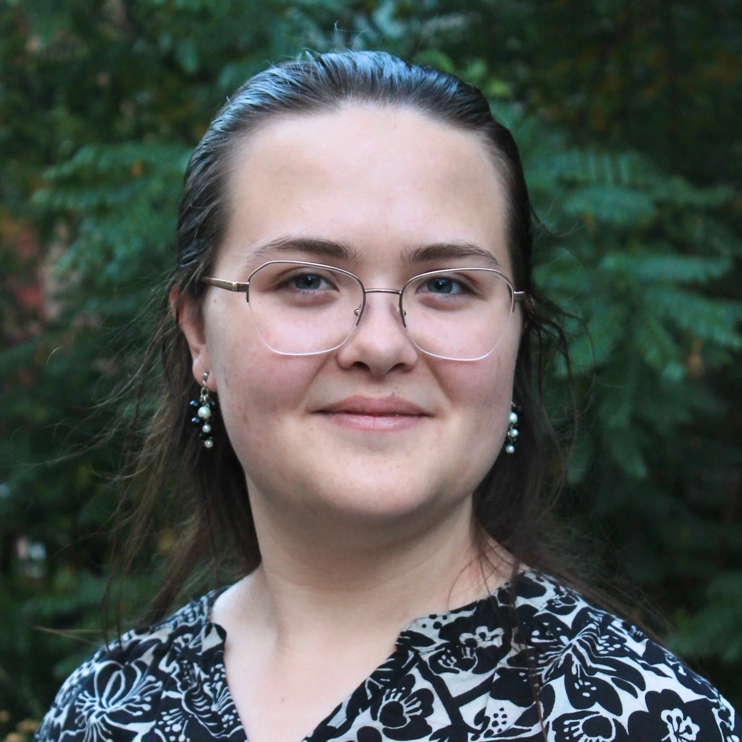 Heather, a light-skinned white-Japanese woman in her 20s, smiles at the viewer. She has long straight brown hair, blue eyes, and gold-rimmed glasses.