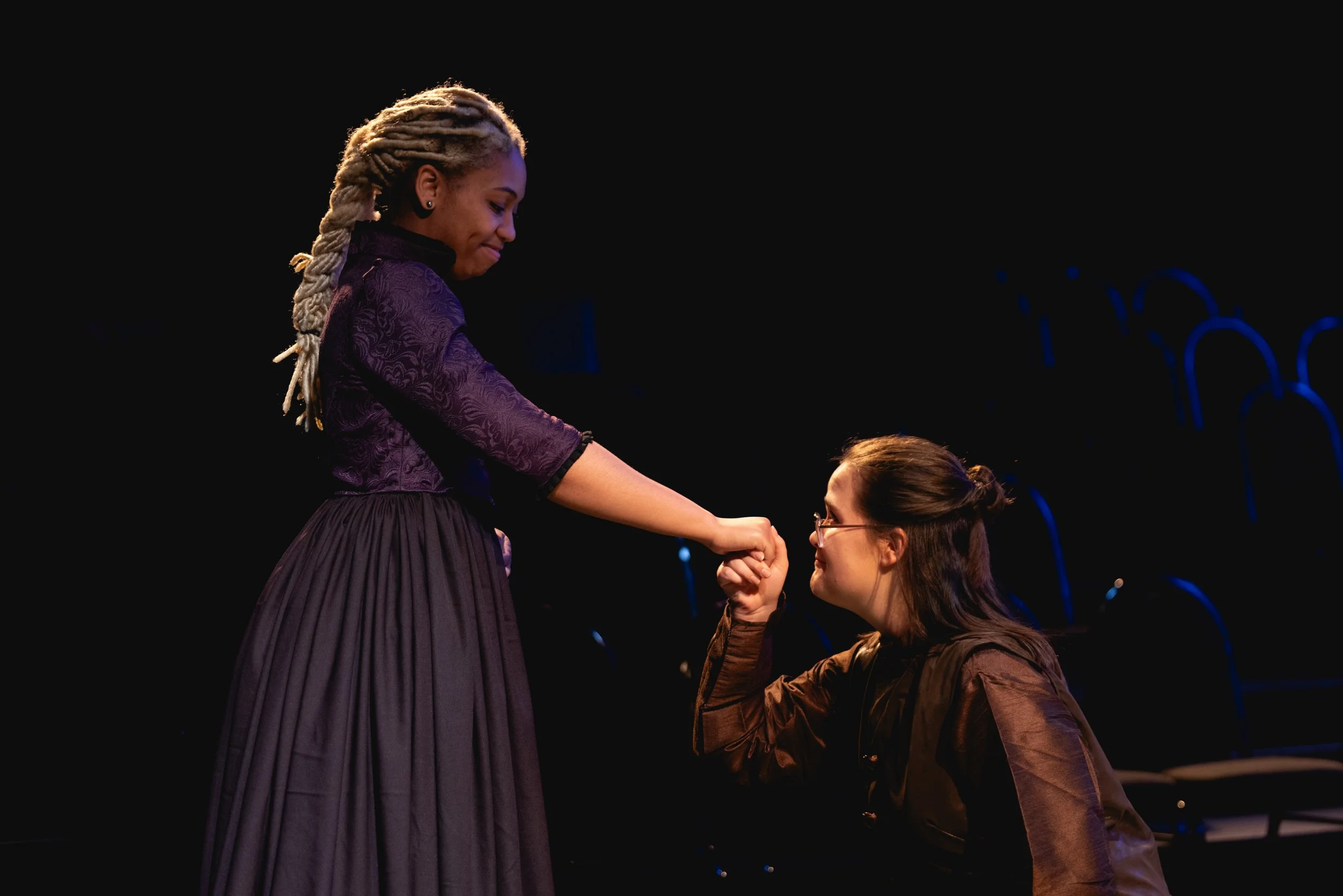 Heather is onstage in a brown buttoned tunic. She kneels and holds the hand of a Black actor with blond braids and a purple dress. The two smile tenderly at each other.