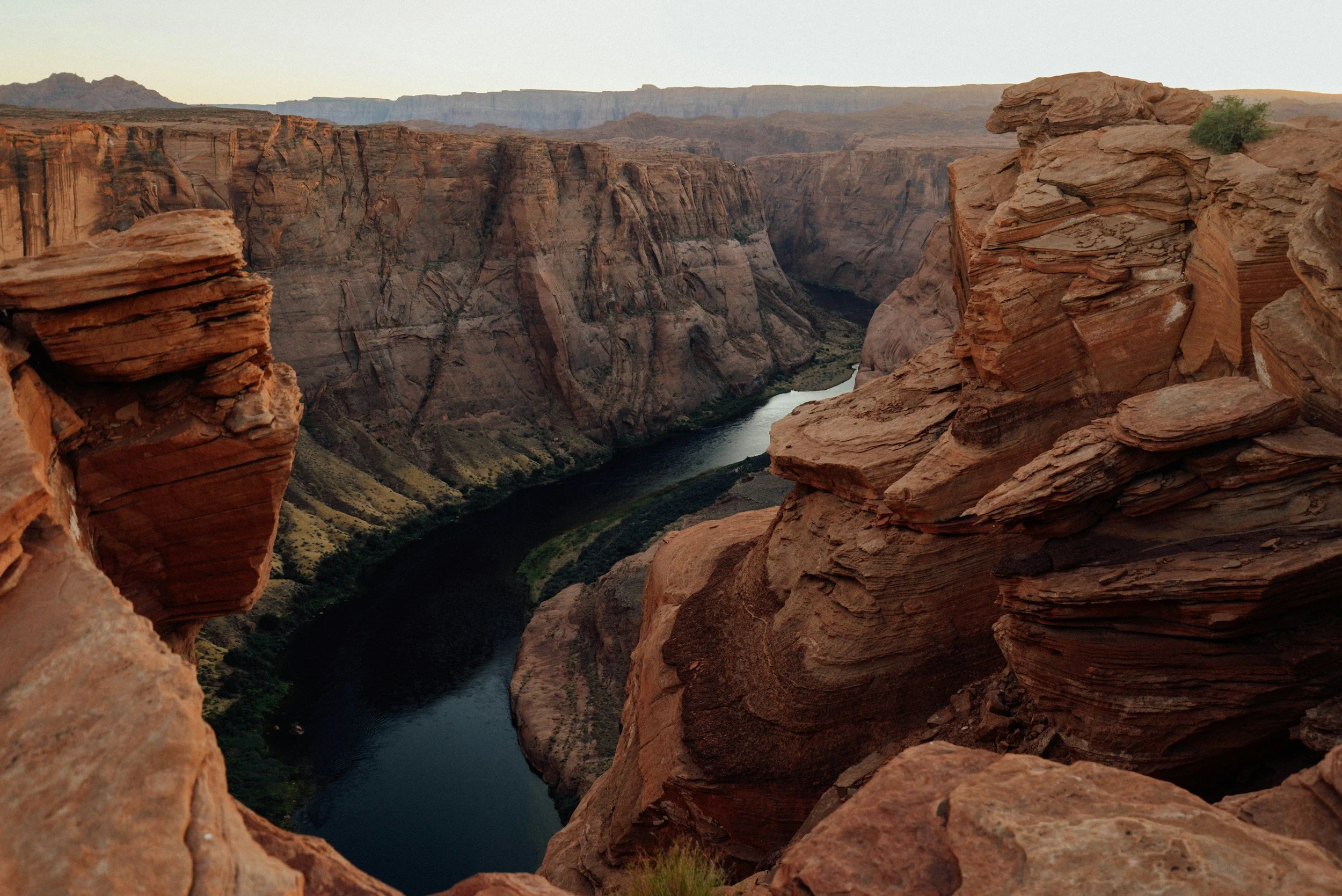 View of a deep canyon with a river flowing at the bottom, surrounded by reddish-brown rock cliffs and rugged terrain, under a clear sky.