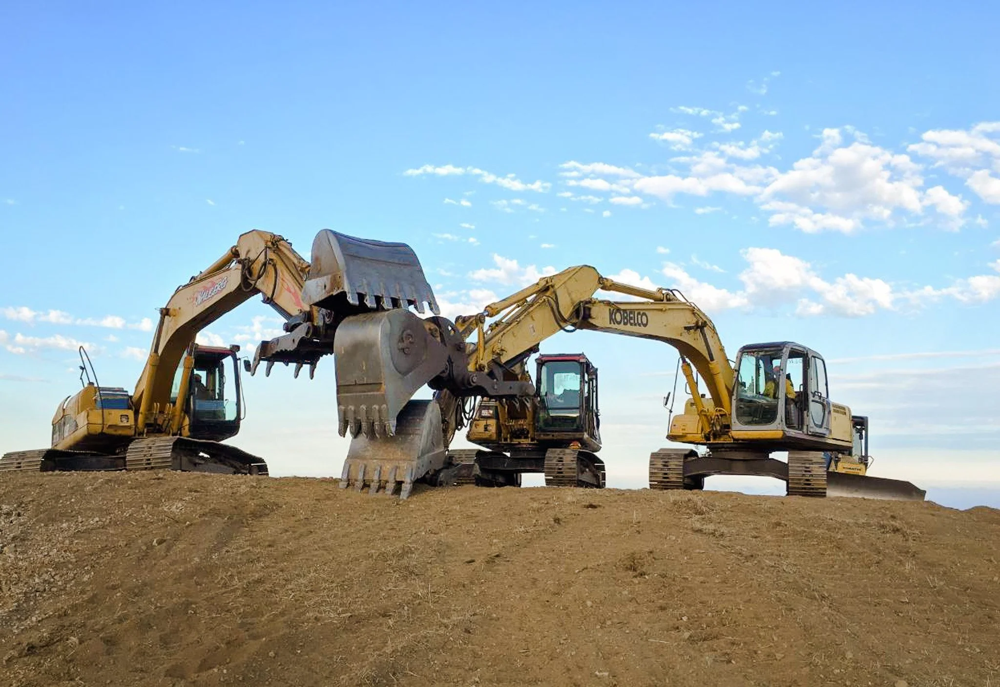 Three yellow excavators with tracked wheels are positioned on a dirt surface under a blue sky with scattered clouds, ready for construction or excavation work.