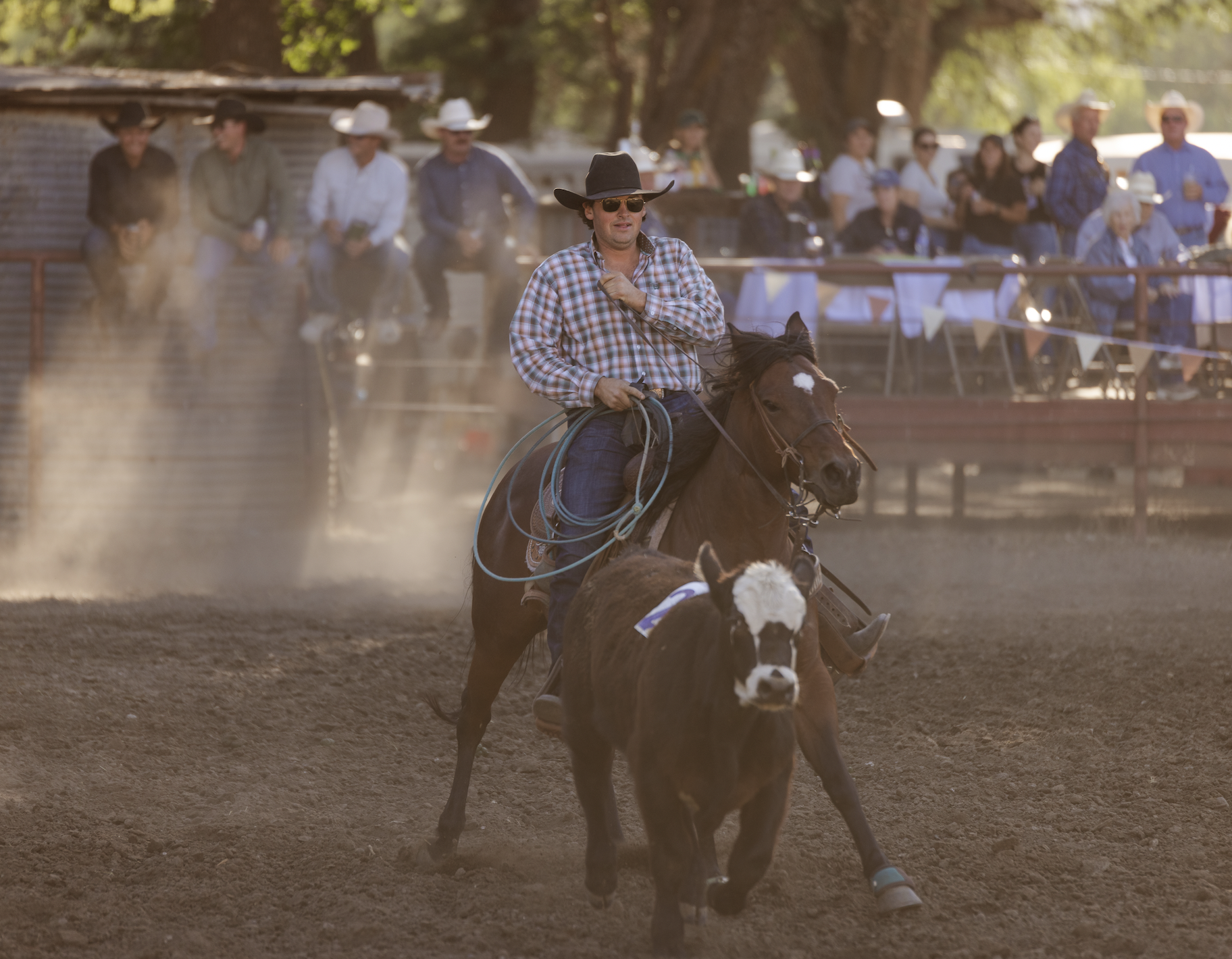 A man in a cowboy hat and sunglasses riding a horse at a rodeo. People are watching behind a fence in the background.