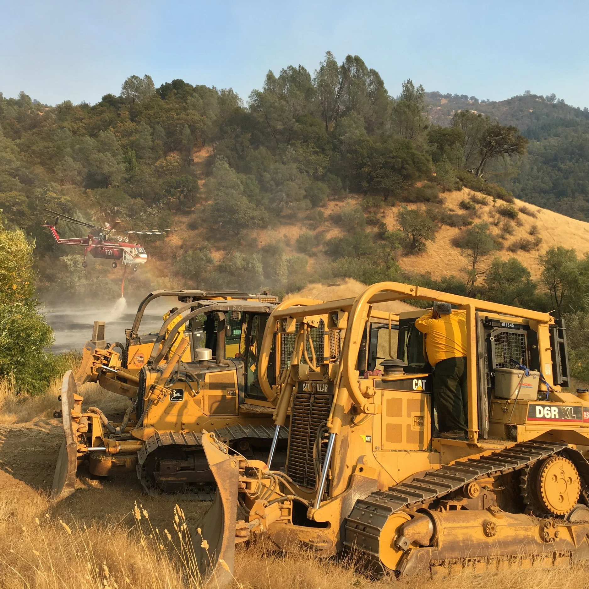 A yellow Caterpillar bulldozer working on a hillside with trees and mountains in the background, and a person in a yellow shirt near the machinery.