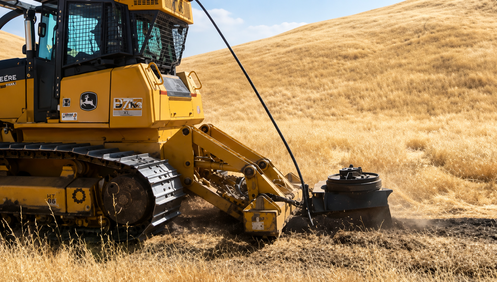 A yellow bulldozer actively working on a dry, grassy field with rolling hills in the background.
