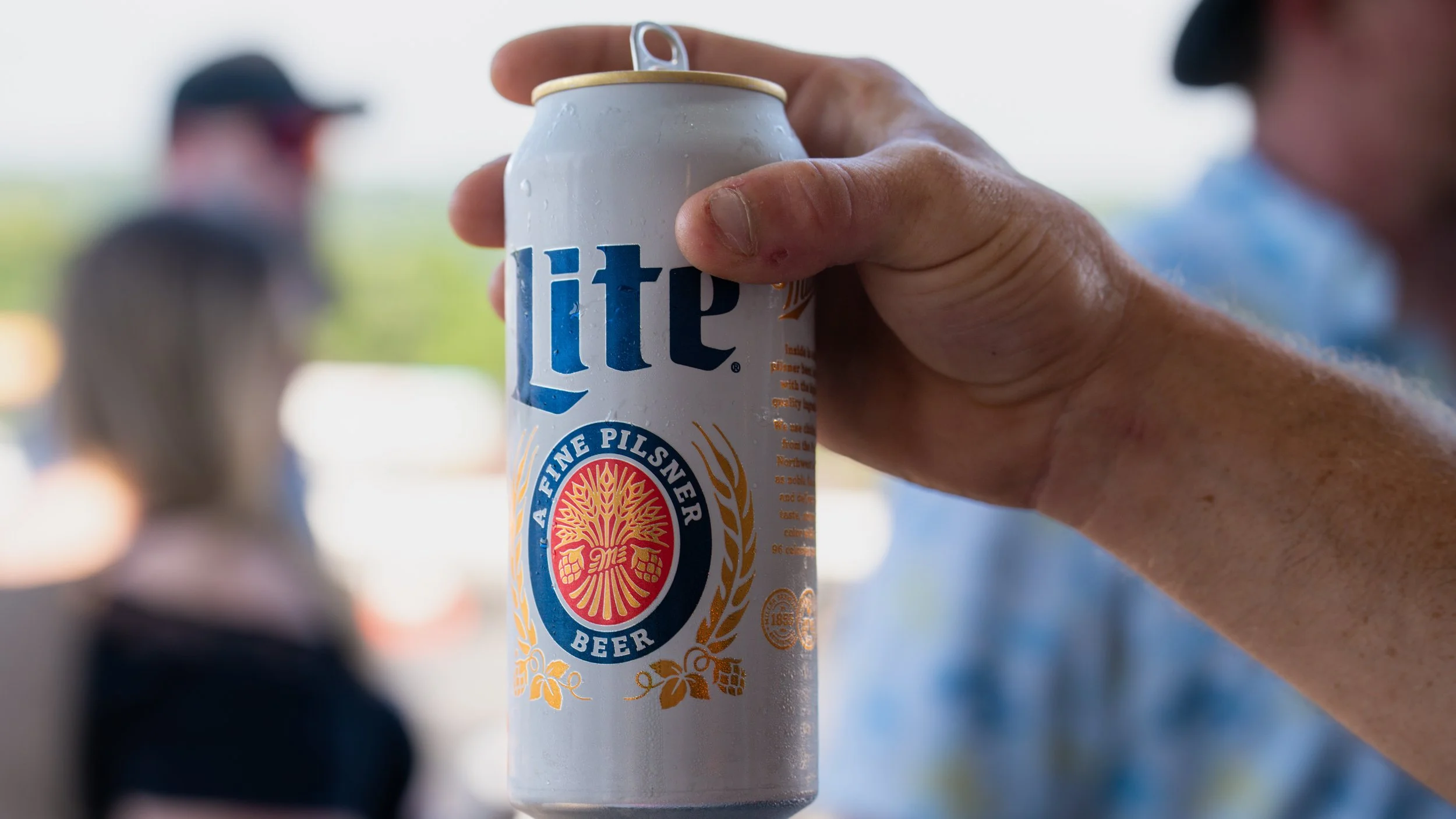 Close-up of a hand holding a white can of Miller Lite beer with an American flag background blurred.