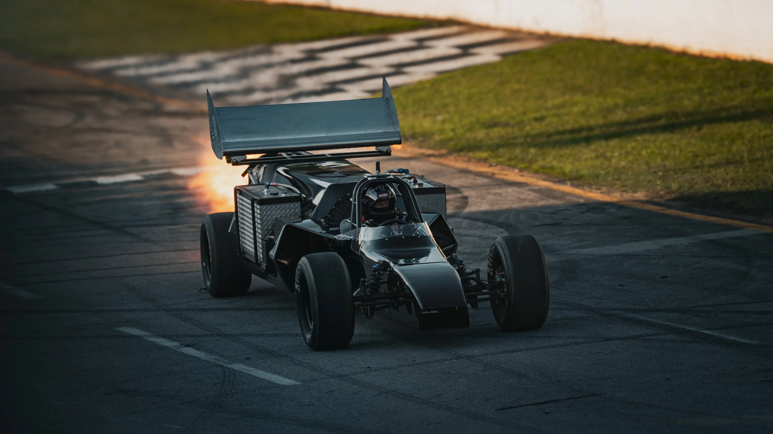 A black racing car with a large rear wing, racing helmet inside, on a race track during a drift or burnout with sparks and smoke underneath, at sunset.