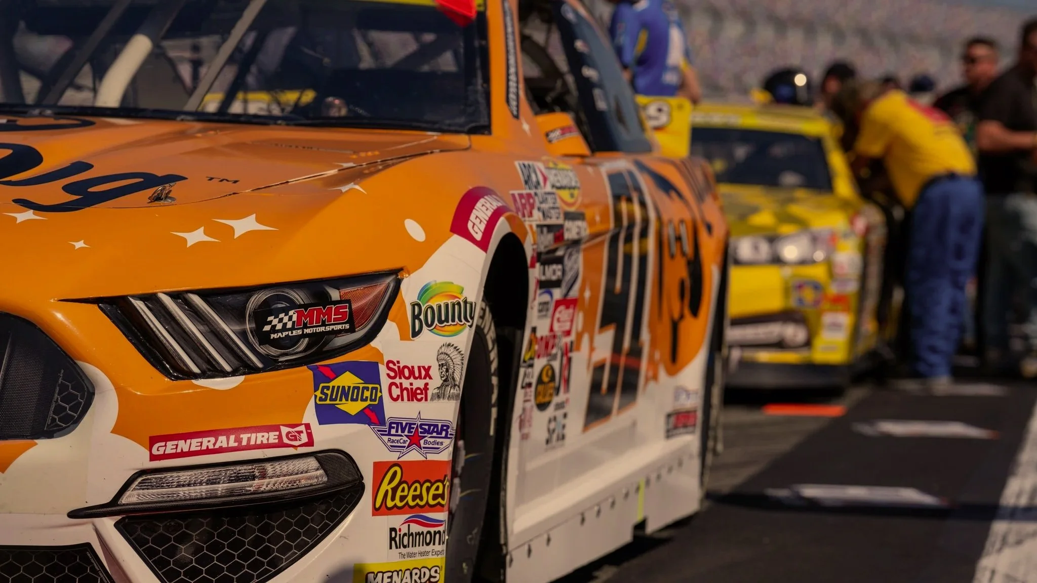 Close-up of a race car with a bright orange and white paint job, featuring various sponsor logos, parked on a racetrack with team members and other race cars in the background.