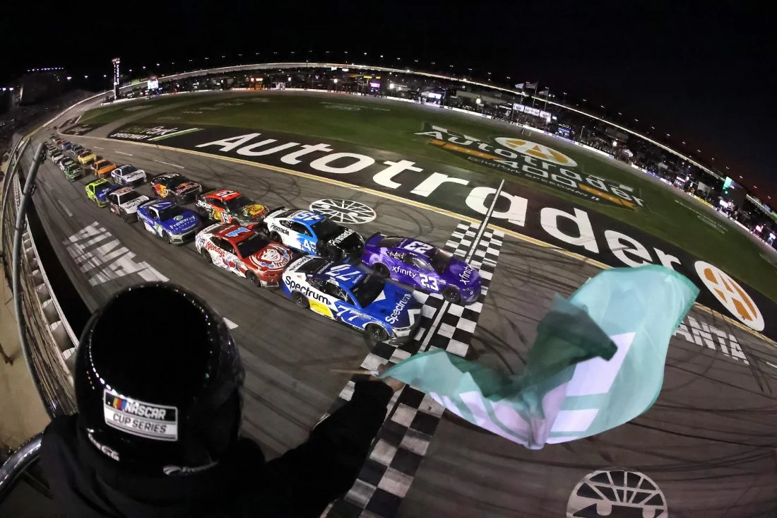 Race cars crossing the finish line at a nighttime NASCAR race, with a person in a NASCAR helmet waving a green flag from a platform