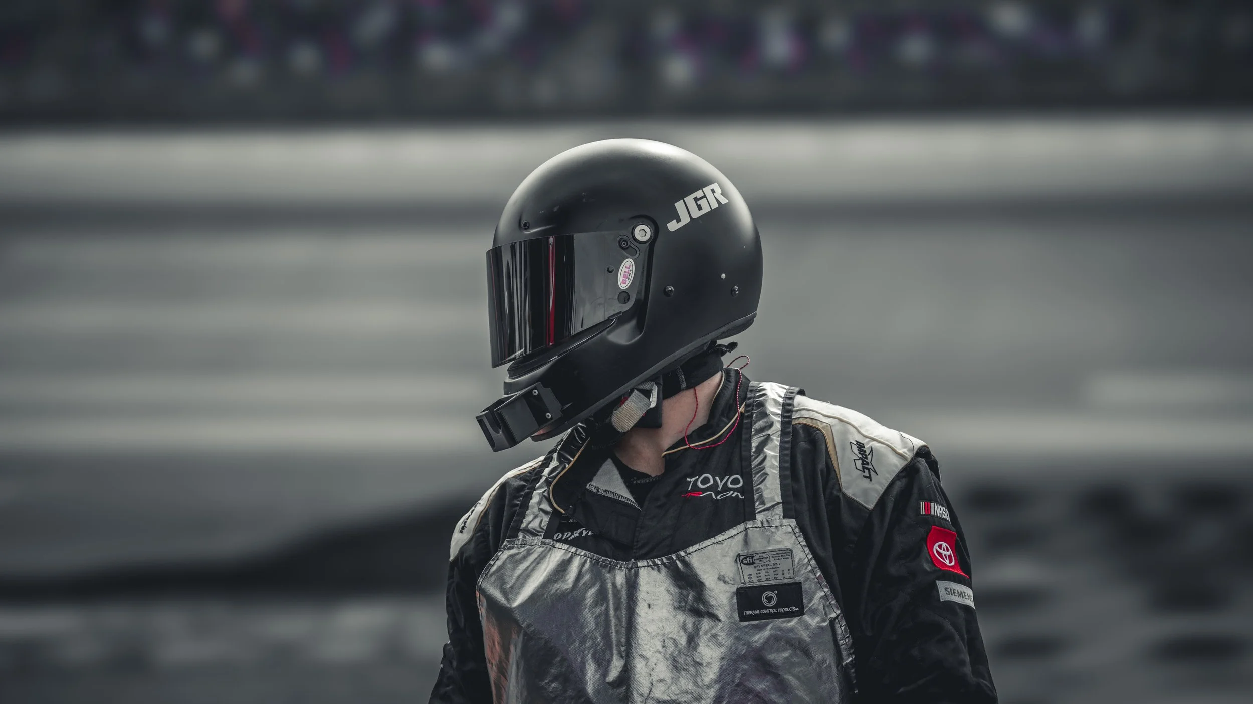 Race car driver wearing a black helmet with a reflective visor and racing suit with sponsor patches, standing on a racetrack.