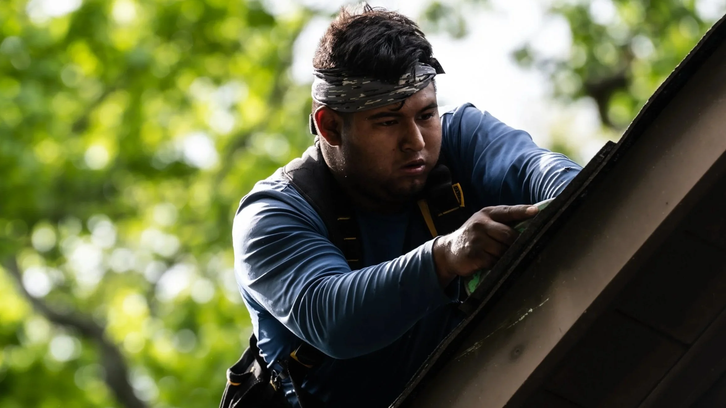 A man wearing a bandana and outdoor gear is working on a roof, surrounded by green trees.