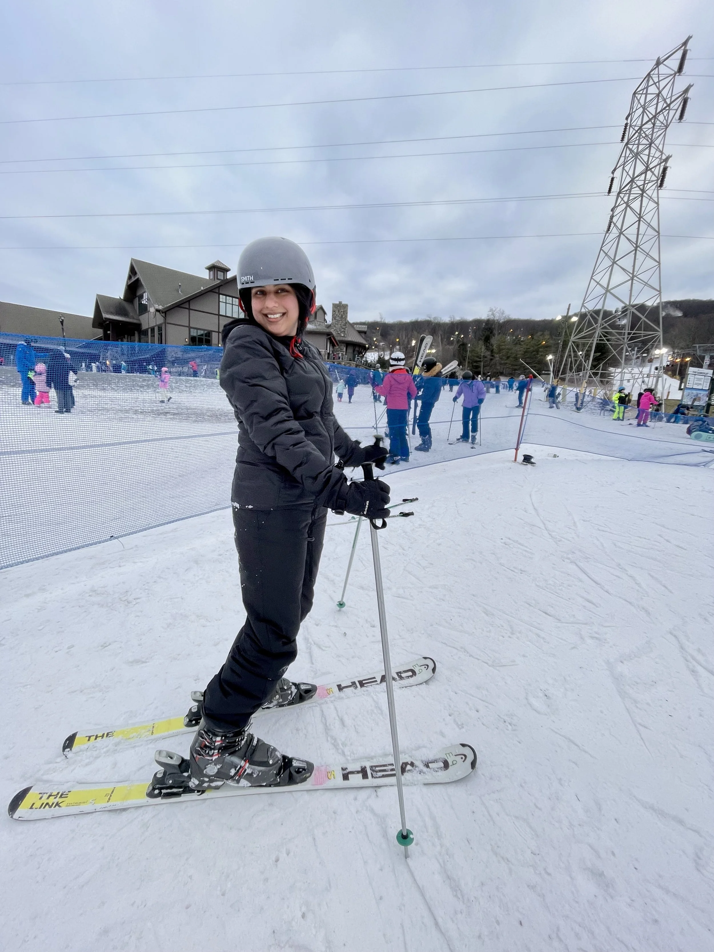 Akanksha Rathod, Founder of Nee's Physical Therapy Hoboken, in ski gear and a helmet standing on skis in a snowy ski resort area.