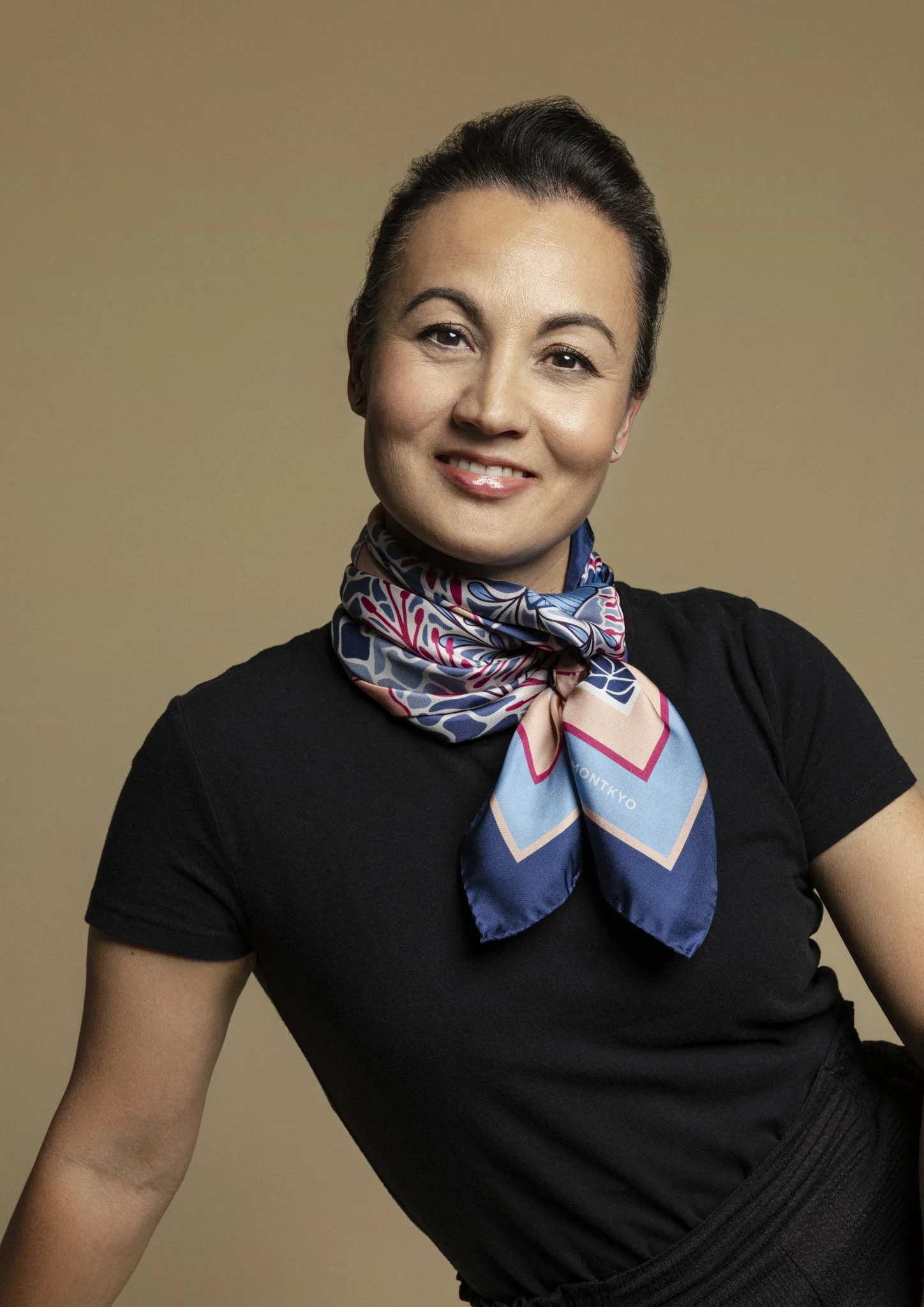 A woman with dark hair tied back, wearing a black top and a colorful patterned silk scarf, smiling against a neutral background.
