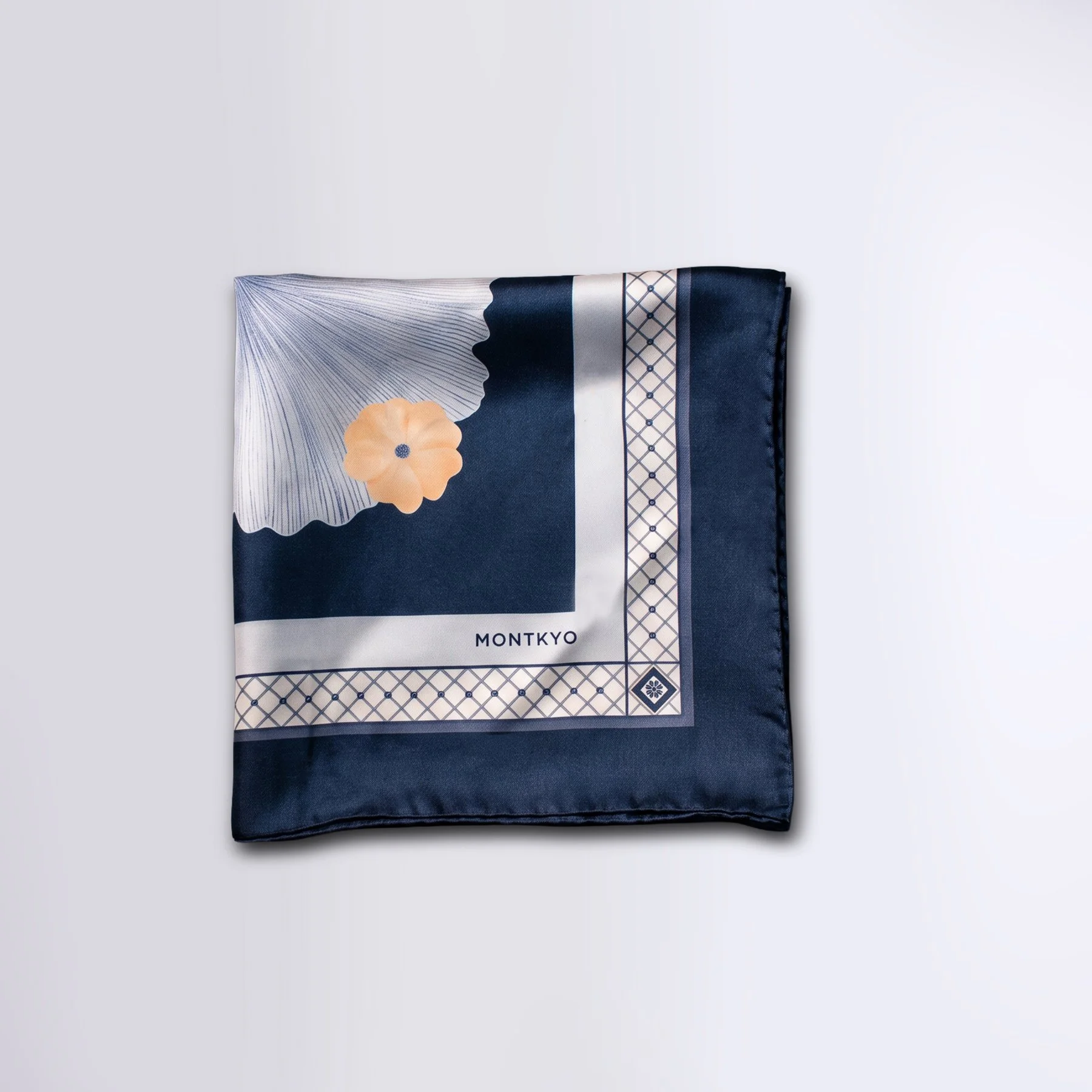 Blue Wings silk scarf by MONTKYO folded into a square and laid flat on a white tabletop, showing a ginkgo leaf and orange flower as part of the scarf design.