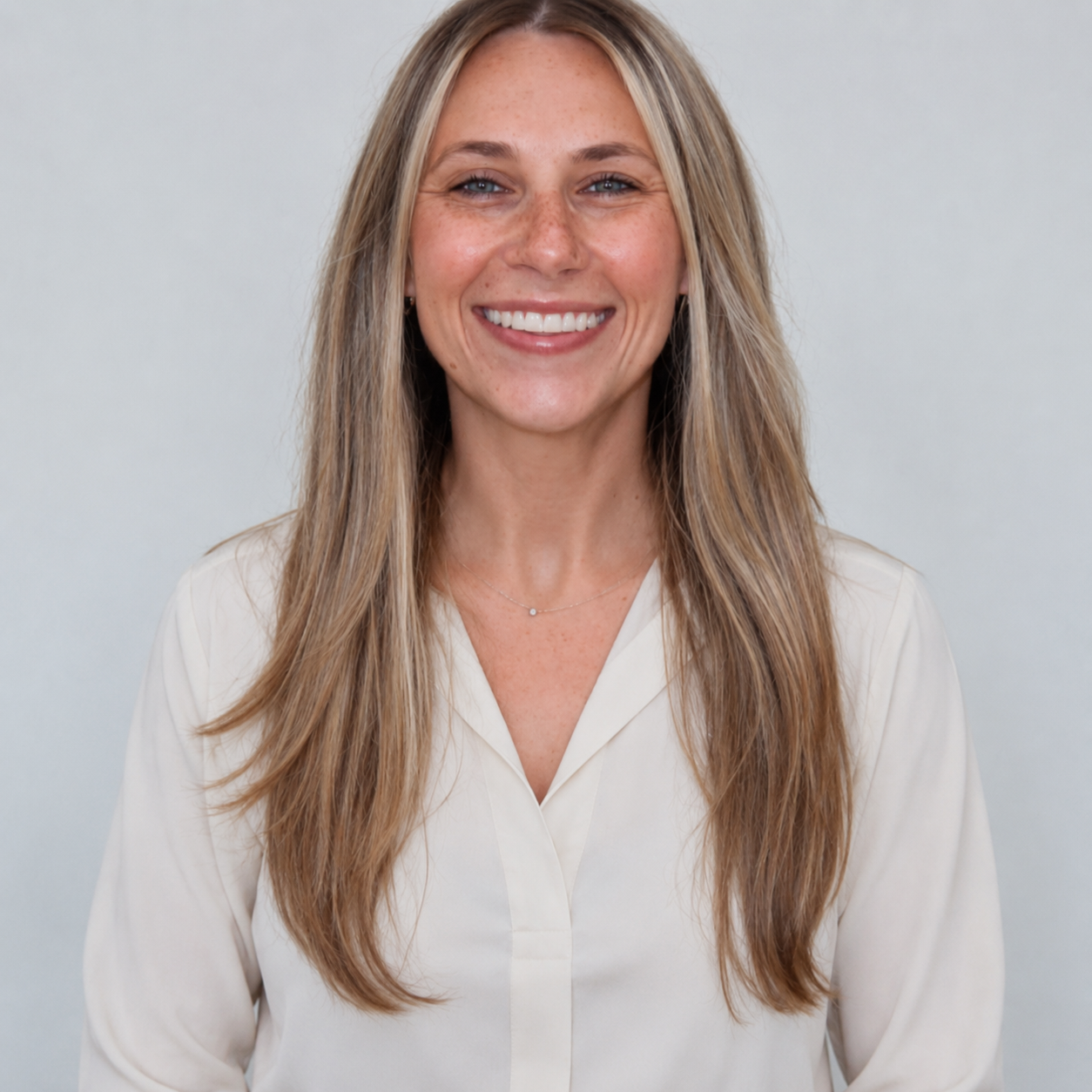 Portrait of a smiling woman with long blonde hair wearing a white blouse and a delicate necklace against a light background.