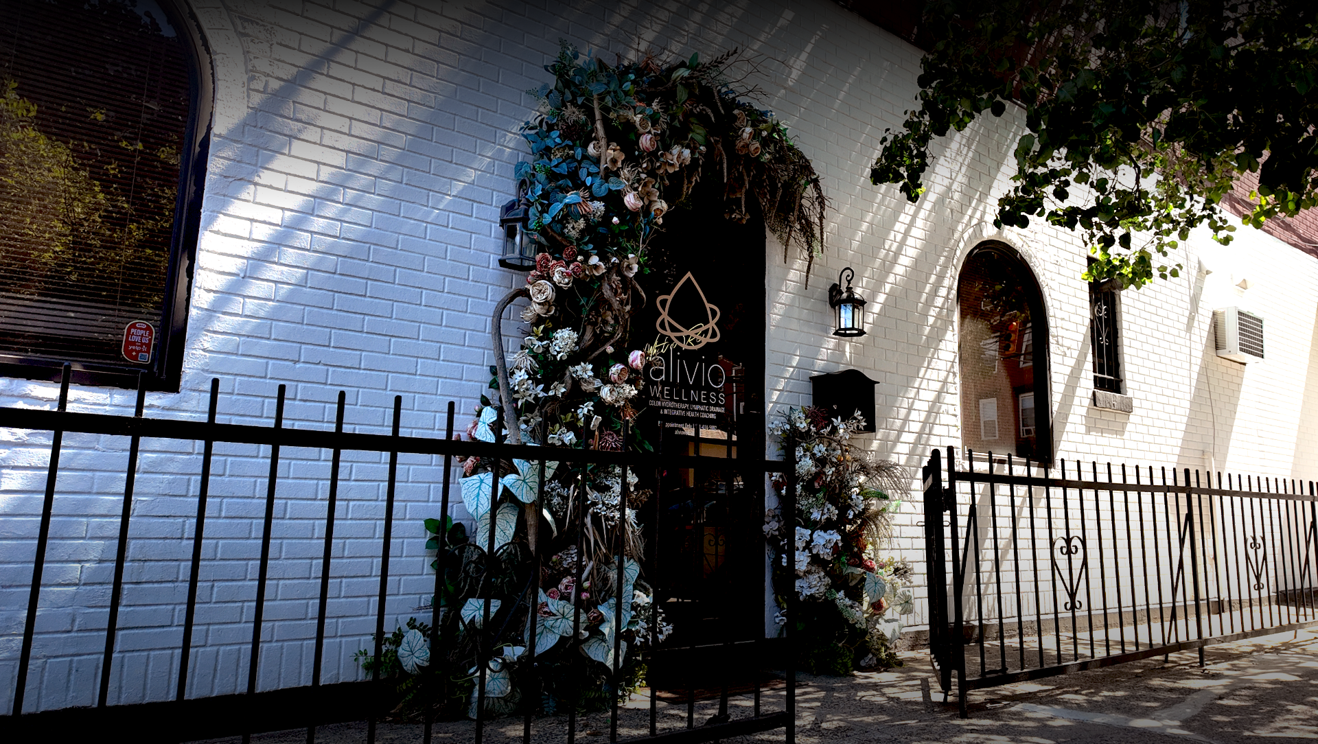Exterior of a wellness center with a white brick wall, a black wrought-iron fence, and decorative floral arrangements around the doorway. Sign reads 'alivio wellness' with smaller text about health treatments.