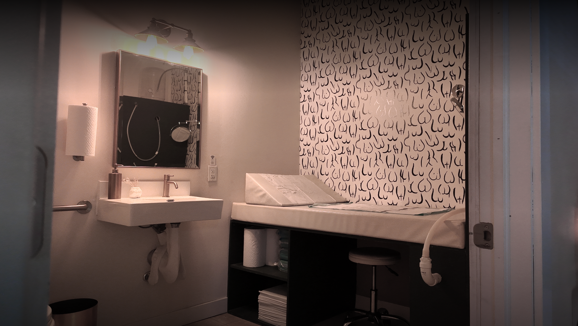 A room with a small white sink, a mirror, a wall-mounted light fixture, a paper towel holder, and a bed with a mattress and a pillow. There is a patterned wall behind the bed and a black shelf underneath it with rolled-up towels and papers.