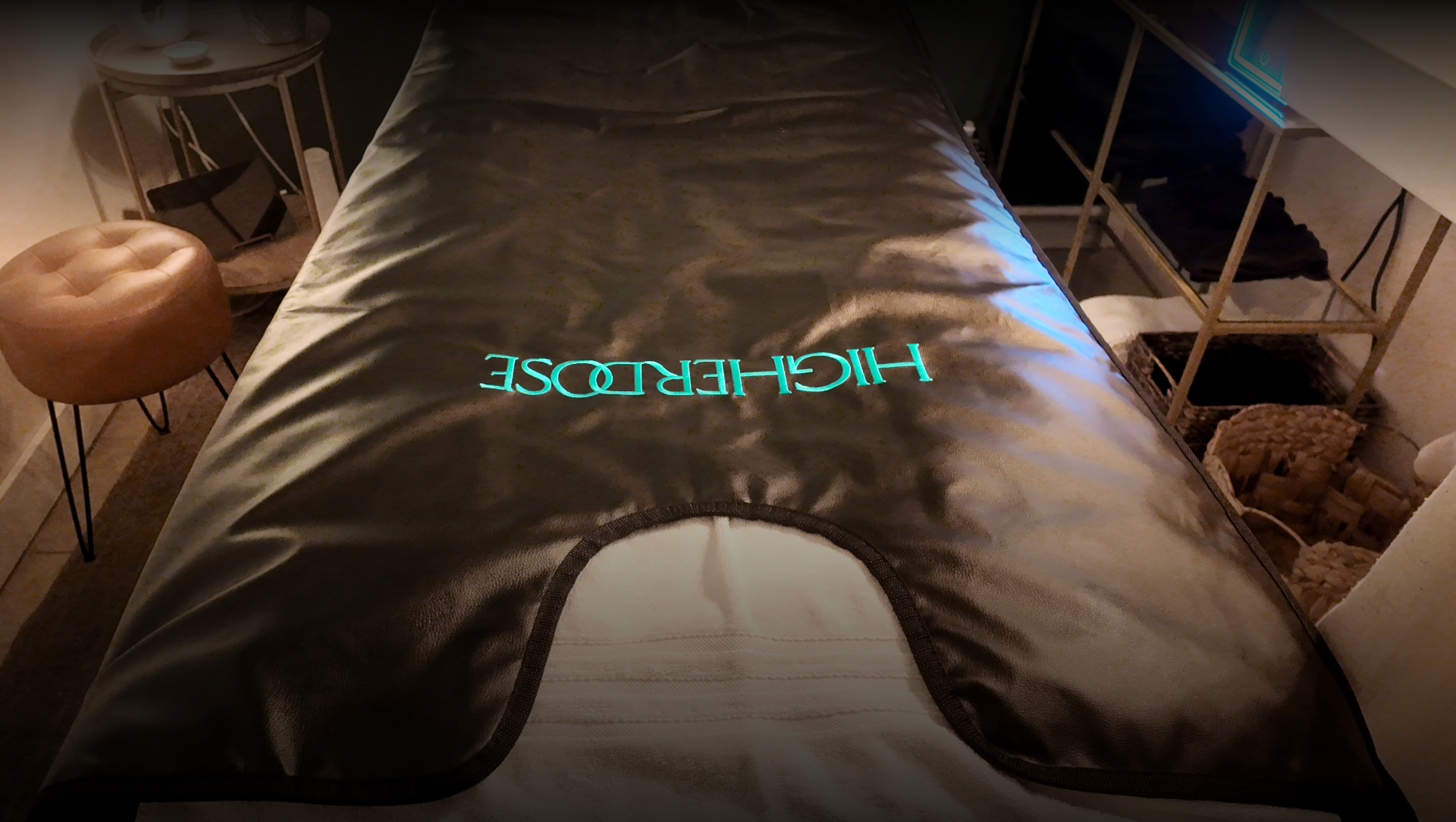 A massage table with the word 'HIGHEREDGE' illuminated on it, surrounded by side tables, a stool, and storage baskets in a dimly lit room.
