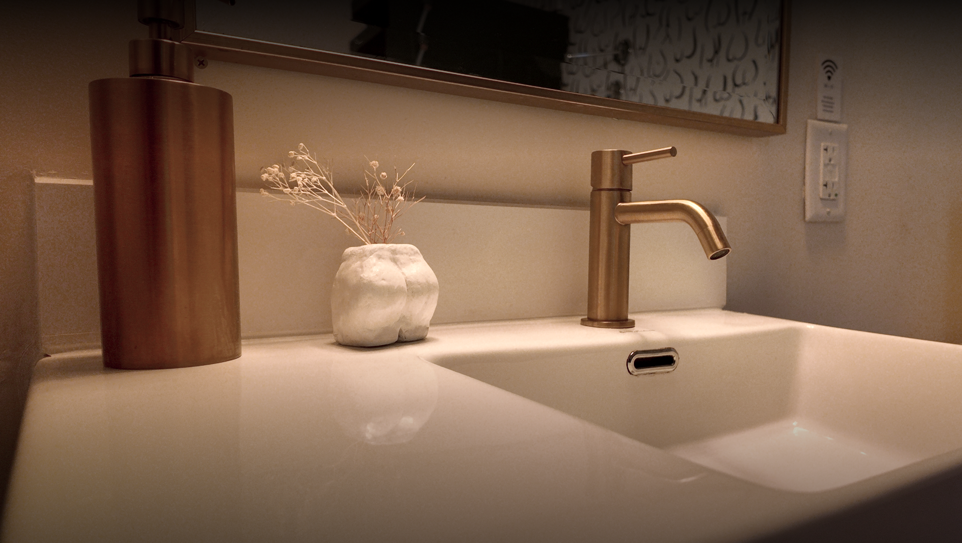 Bathroom sink with a gold faucet, a white textured vase with dried flowers, a copper-colored soap dispenser, a mirror, a power outlet, and a wall with a Wi-Fi symbol.
