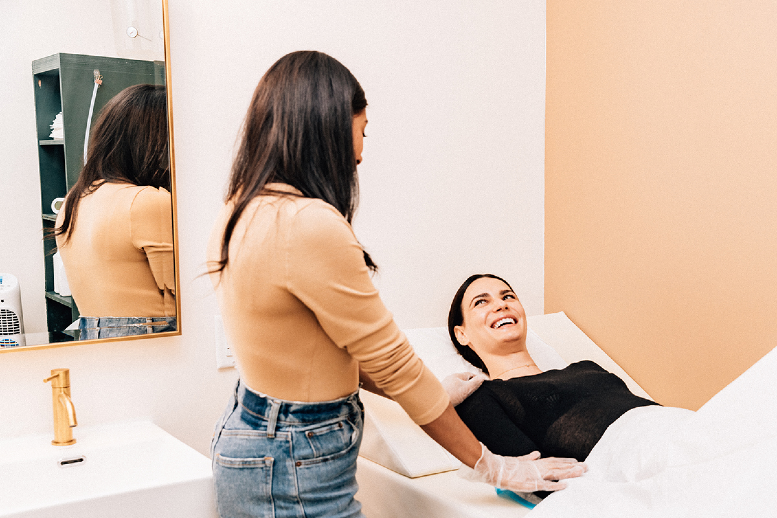 A woman with long dark hair lying on a white medical examination bed, smiling and looking up. Another woman with long dark hair and wearing a beige top and blue jeans is standing next to her, looking at her. The setting appears to be a medical or wellness clinic with a mirror on the wall, a small shelf with items, and a gold faucet on the sink.