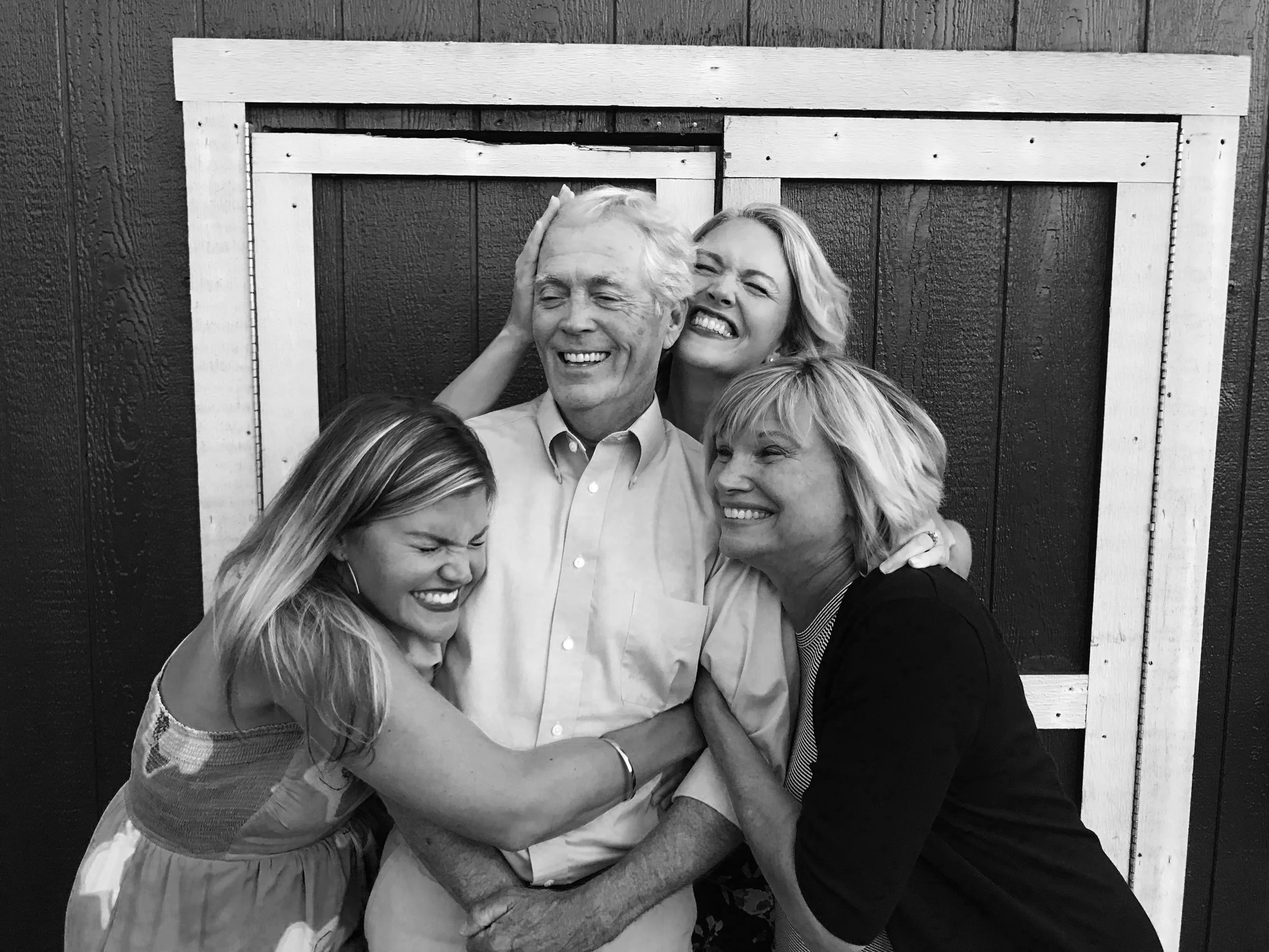 Tom Connelly is surrounded by his daughters, Kelly and Katie, and his wife, Wendy. The ladies are all hugging Tom and they are standing in front of a barn door.