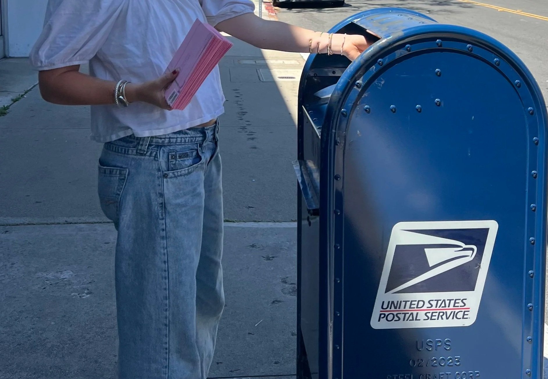 A person depositing a letter into a blue USPS mailbox on a city sidewalk. The person is wearing a white top, light blue jeans, and jewelry on the wrist.