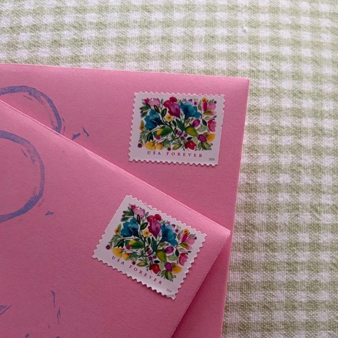 Close-up of a pink greeting card with two colorful postage stamps featuring a floral design, placed on a checkered fabric surface.