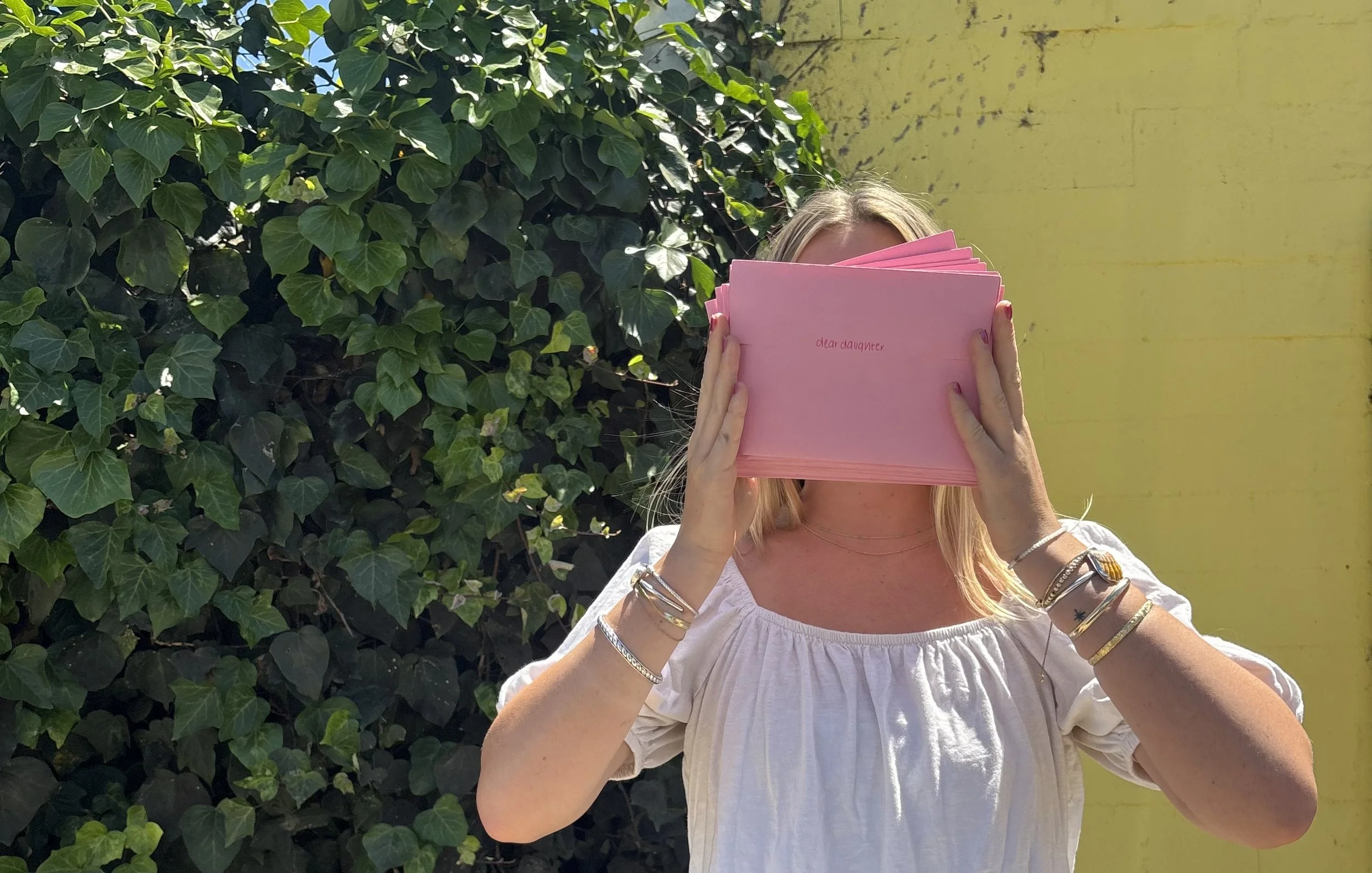 Woman with blonde hair holding a pink greeting card in front of her face while standing outdoors with green leafy bushes and a yellow wall in the background.