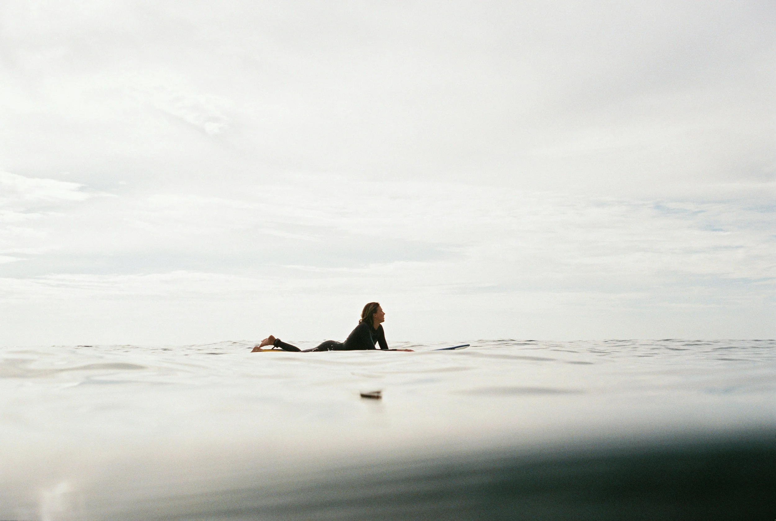 A woman in a black wetsuit is lying on a surfboard in the ocean, looking at the horizon with a cloudy sky overhead.