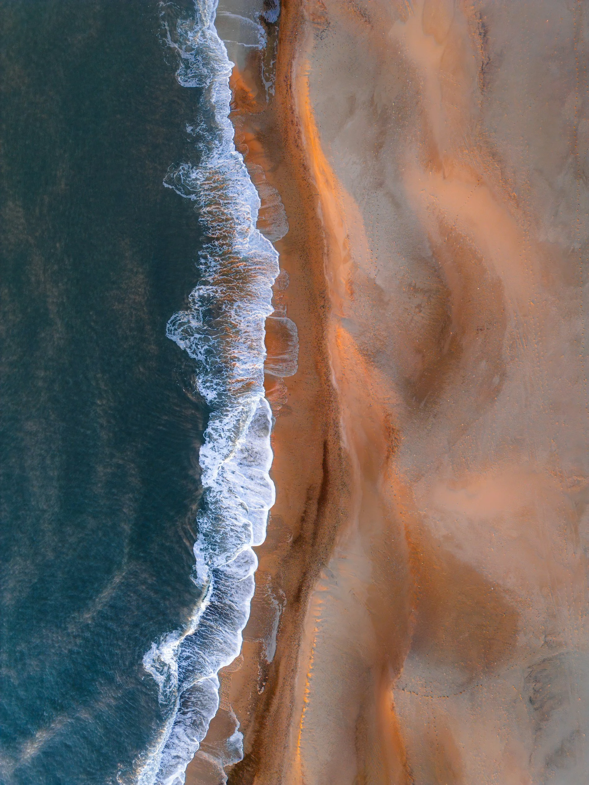 Aerial view of coastline showing waves crashing onto sandy beach at sunset.