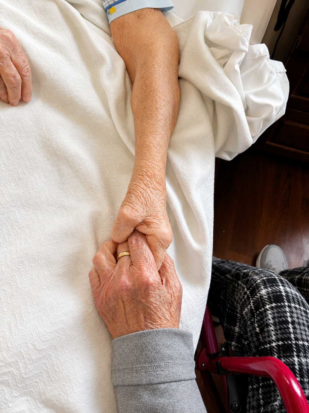 Close-up of an elderly person holding hands with a caregiver or family member, lying in a hospital bed.