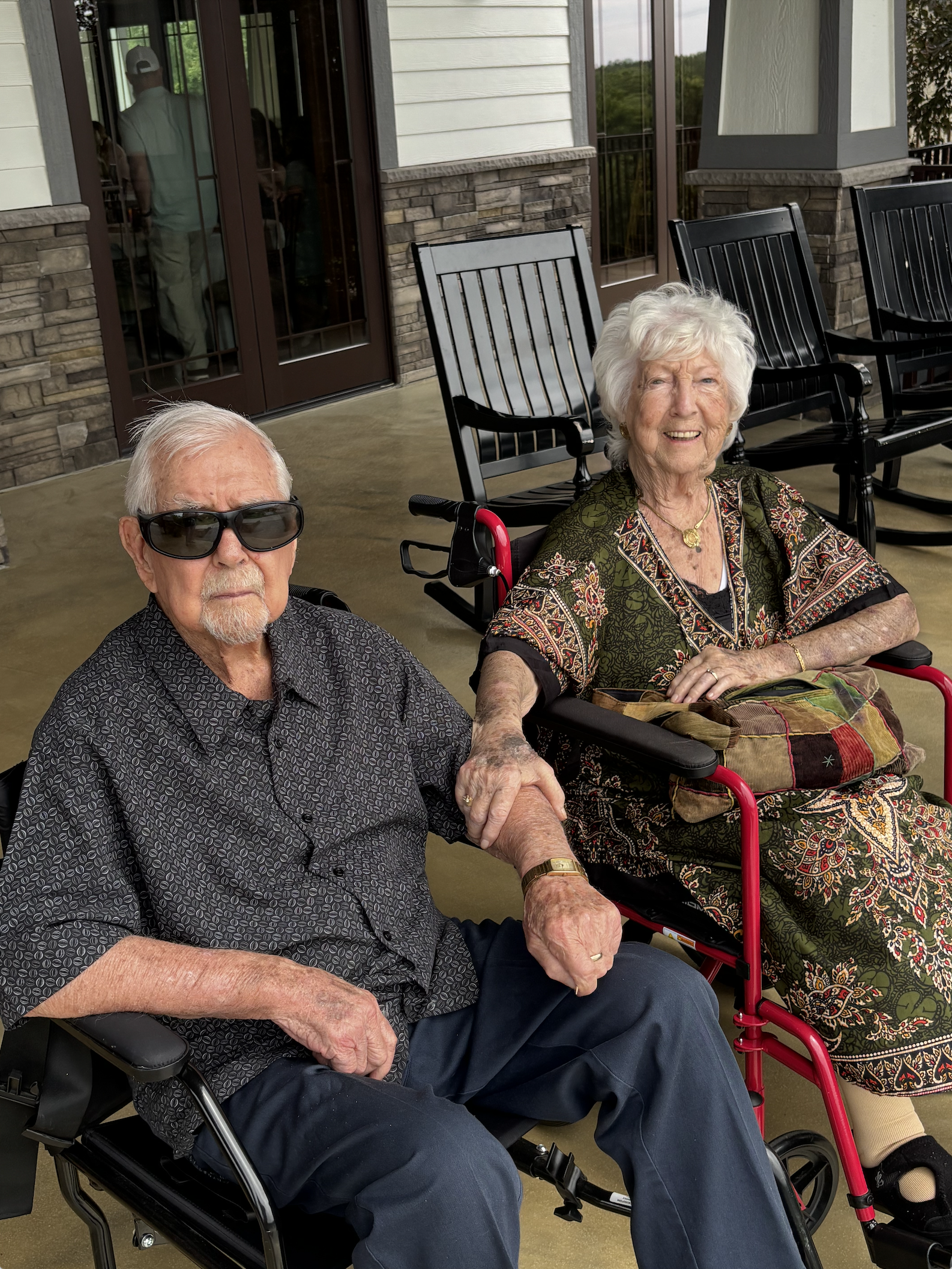 An elderly man with white hair and sunglasses sitting in a wheelchair, holding hands with an elderly woman with white hair and a patterned dress, sitting in a wheelchair on a porch with black rocking chairs and a stone and siding house in the background.
