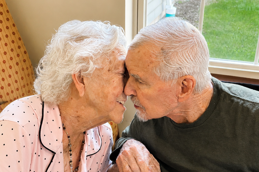 An elderly woman and an elderly man are touching foreheads and smiling with their eyes closed inside a room with a window overlooking a grassy yard.