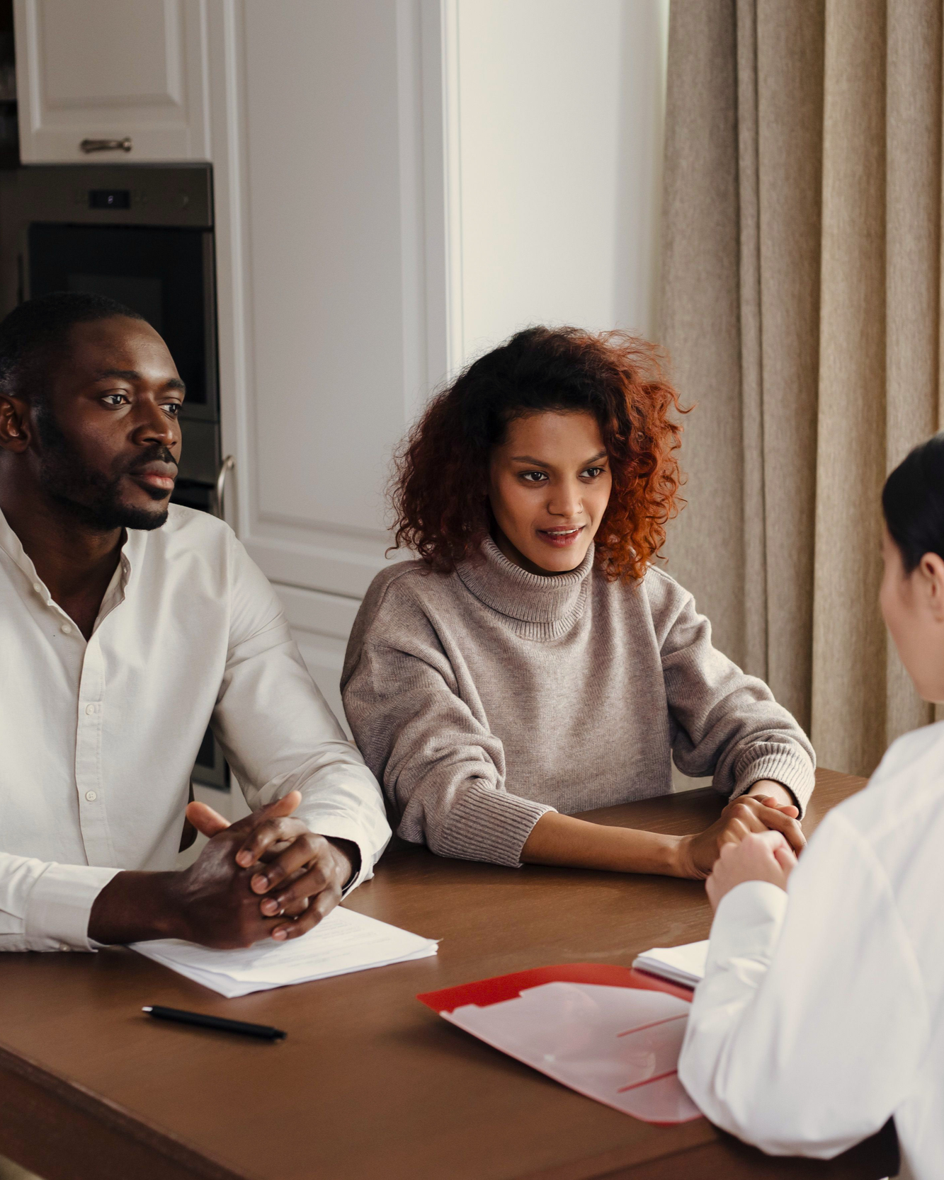 Two men and a woman having a meeting with a person in a white coat at a table with documents and a red folder.