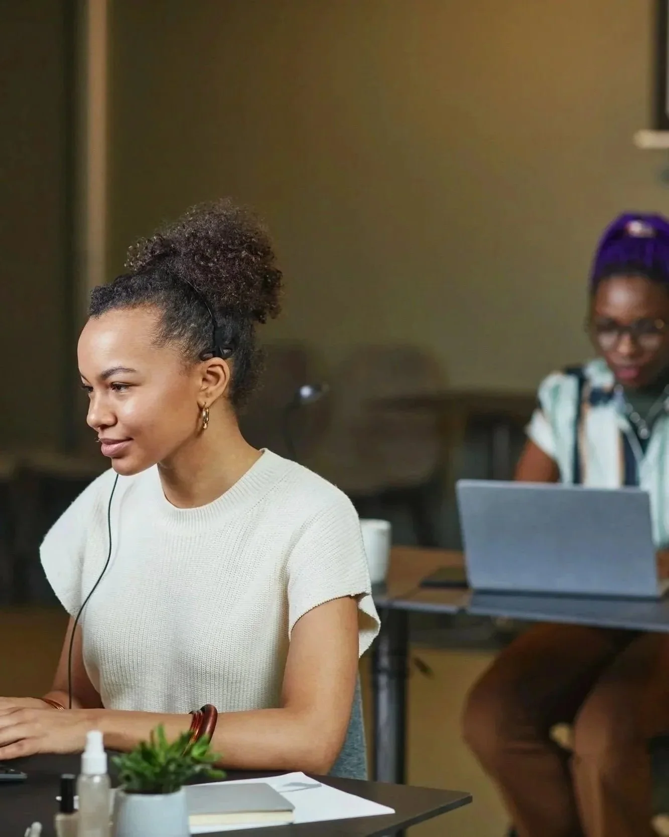 Two women working in an office: one with curly hair in a ponytail, wearing headphones, and the other with her hair in a bun and wearing glasses and a colorful top.